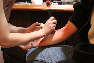 A student's hands apply make up to another student's arm, resembling a rash. 