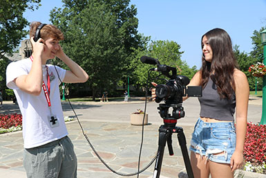Two students smile at one another, one wearing headphones and the other with a hand on a camera and tripod.