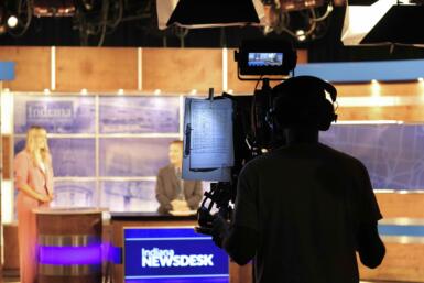 Person operates a TV camera with two people behind a news desk.