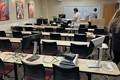 Rows of tables stand in a classroom holding new computer monitors and technology. A person stands in the background. 