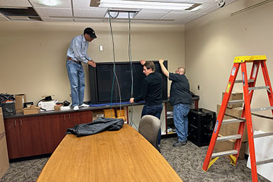 Three men hold onto a TV screen in the BFCA's seminar room. Wires hang from the ceiling. 