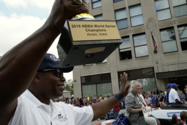 A man holds a trophy and waves from a car.