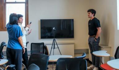 Three students stand in a classroom while one reads from their phone preparing to film another student using a tripod and camera standing in between the people.