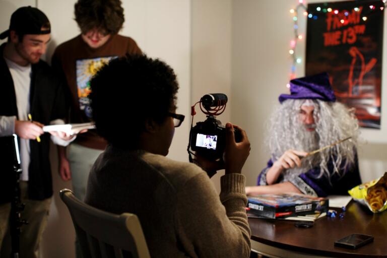 Person dressed up in a wizard costume points a wand at a person filming them while two other people stand to the side overlooking a piece of paper.