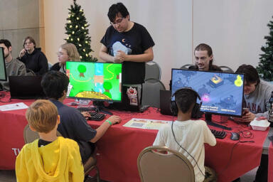 Game design students sit behind a long table and watch children test their game projects on computers.