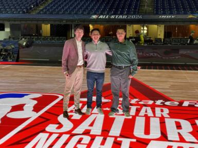 Three students stand in the middle of a basketball court that displays the NBA logo.