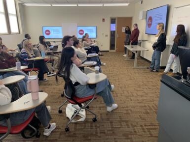 Four students stand at the front of a classroom presenting to several other students in chairs. 