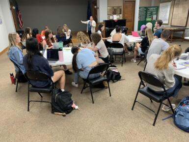 Students sit at tables, listening and taking notes as a speaker talks. 