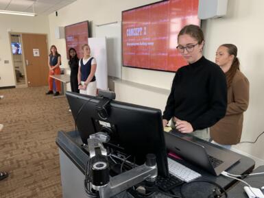 Students stand at the front of a classroom with a presentation pulled up on screen.