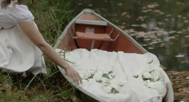 A woman lays her hand on a white blanket inside a rowboat by the water. 