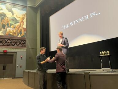 Two students hold a trophy while a third watches them from a stage. 