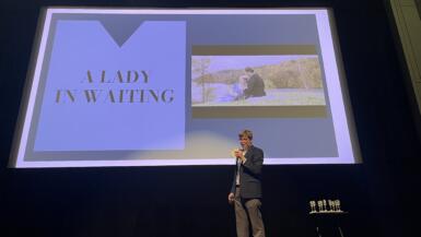 A man on a stage stand in front of a screen that shows a student short film thumbnail with the title "A Lady in Waiting."