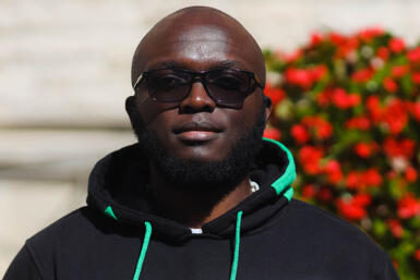 A headshot of a man in sunglasses and a hoodie with greenery behind him.