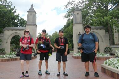 Four people with camera equipment strapped around their backs and necks stand on the brick sidewalk outside in front of the Sample Gates at IU Bloomington campus.