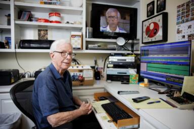 Person sits at a desk with a computer and keyboard in front of them, several recording equipment stacked, and a TV mounted on the wall.