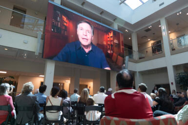 Video of Bob Costas plays on the screen in the media school commons.