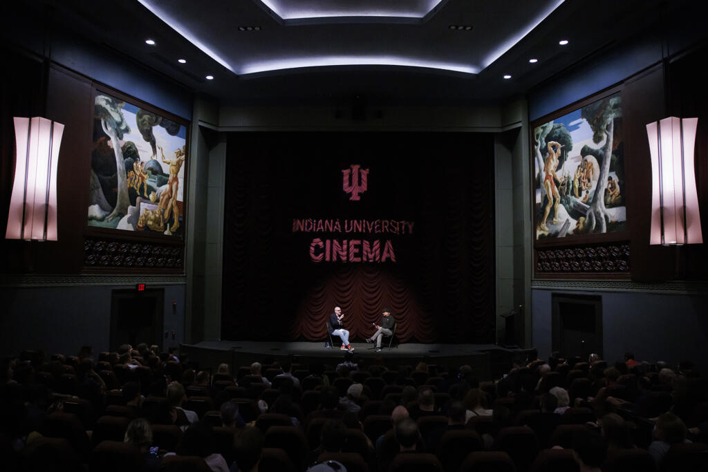 A theater with two men sitting in chairs on stage. An audience watches. "IU Cinema" is on the curtain behind them.