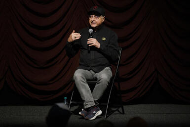 Michael Uslan speaks into a microphone while sitting in a chair onstage.