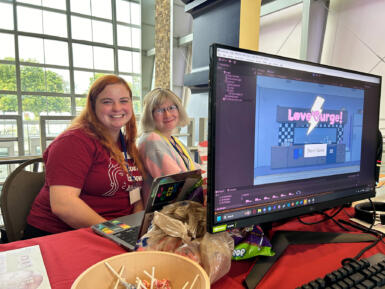 Two women smile behind a computer monitor with a game title screen pulled up reading, "Love Surge."