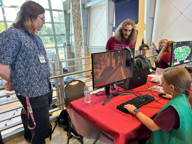 A child uses a mouse and keyboard to play a game on a computer monitor while two Media School students watch.