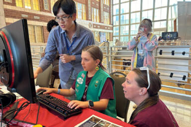 Two adults stand around a young girl as she sits in front of a computer monitor and uses a mouse and keyboard. In the background, another person is taking a photo with their phone.