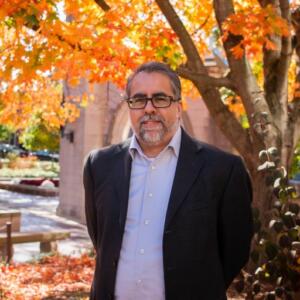 Headshot of Gerry Lanosga standing outside near the Sample Gates with autumn leaves on the trees behind him.