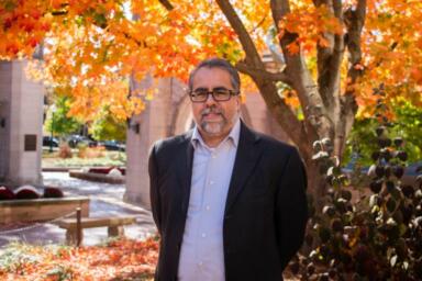 Headshot of Gerry Lanosga standing outside near the Sample Gates with autumn leaves on the trees behind him.