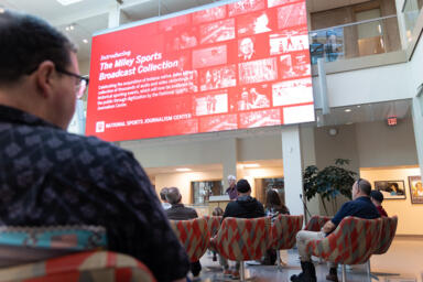 screen displaying the Miley sports journalism collection with John Miley speaking at the podium and people sitting in chairs listening.