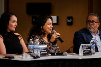 Three people are seated at a table with the person in the middle speaking into a handheld microphone.