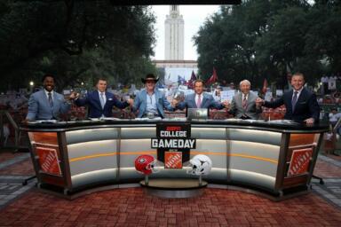 Give people of the College GameDay ESPN program are seated on stage with several people standing behind them.