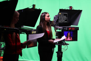 Two people holding scripts stand next to television prompters in front of a green screen.