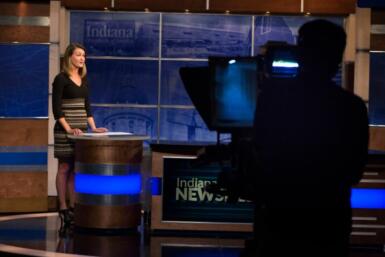 Person stands in a studio behind a desk with people off to the side operating a camera filming the reporter.