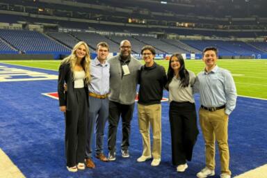 Group of people stand on the Colts football field with an empty stadium behind them.