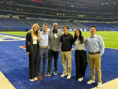 Group of people stand on the Colts football field with an empty stadium behind them.