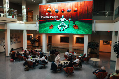 Students sit at tables below a big screen.