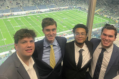 Four people stand in a press box in a football stadium.