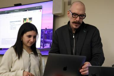 Two people stand in front of a computer with a large projector displayed behind them.