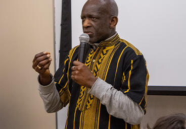 Son of Paulin S. Vieira speaks at his celebrations of life wearing a yellow and black shirt, addressing a room.