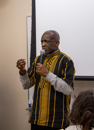 Son of Paulin S. Vieira speaks at his celebrations of life wearing a yellow and black shirt, addressing a room.