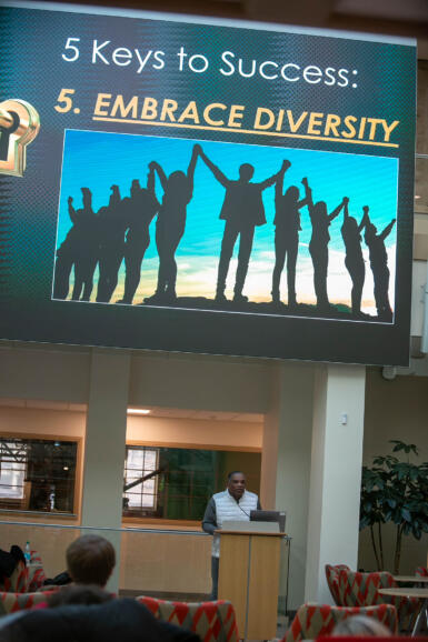 Mark Grant stands at a podium below a screen showing an image of people holding hands with the text, "Embrace Diversity," above it.