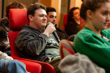 A student holds a microphone while sitting among other students.