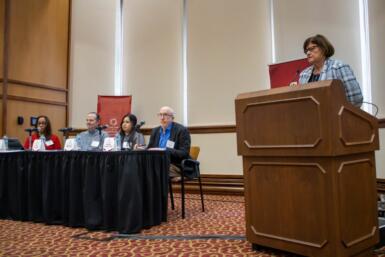 Panelists Tim Evans, Daniela Molina, and Henry Schuster sit on stage with Kathleen Johnston standing in front of a podium to the side of them.