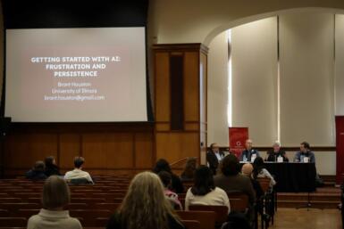 A wide shot of a large projector displaying "Getting Started with AI: Frustration and Persistence" with several people seated in an audience looking toward four people seated on a stage at the front of a large room.