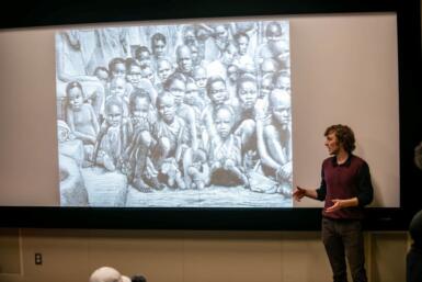 Person stands next to a large screen projecting an image of several children seated, huddled together looking at the camera.