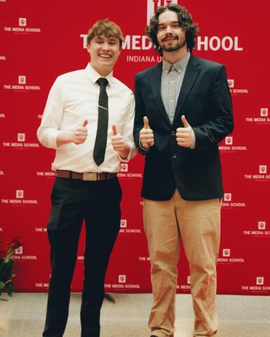 Two people give the camera a thumbs up while standing in front of a Media School backdrop.