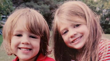 Two children are smiling while seated outside.
