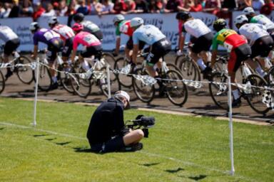 Person sitting on grass with a camera in his hand films as several bicyclists round a corner during the Little 500 race in Bloomington, Indiana.