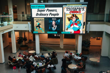 A lot of people seated in a hall watching a big screen with three posters displaying the characters Invincible, Buffy the Vampire Slayer and The Uncanny X-Men, which reads, 
