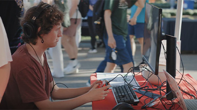 Picture of a man wearing black headphones and holding wires in front of a keyboard on a table, with a crowd in the background.