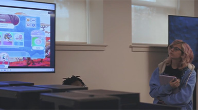 A woman presenting to the class with a screen in the background.